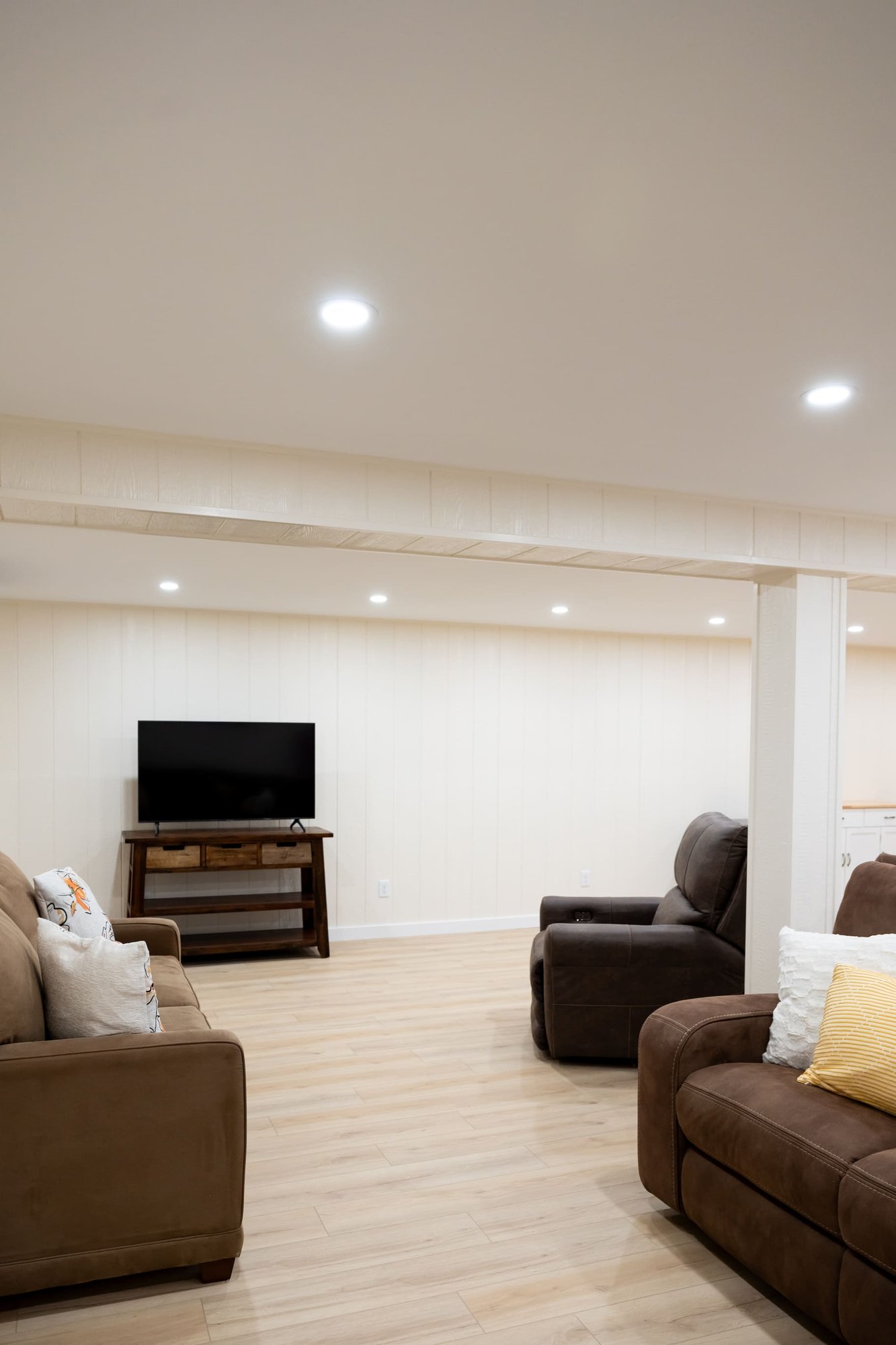 Brown couches, tv on dark wood stand in open white basement remodel with recessed lighting